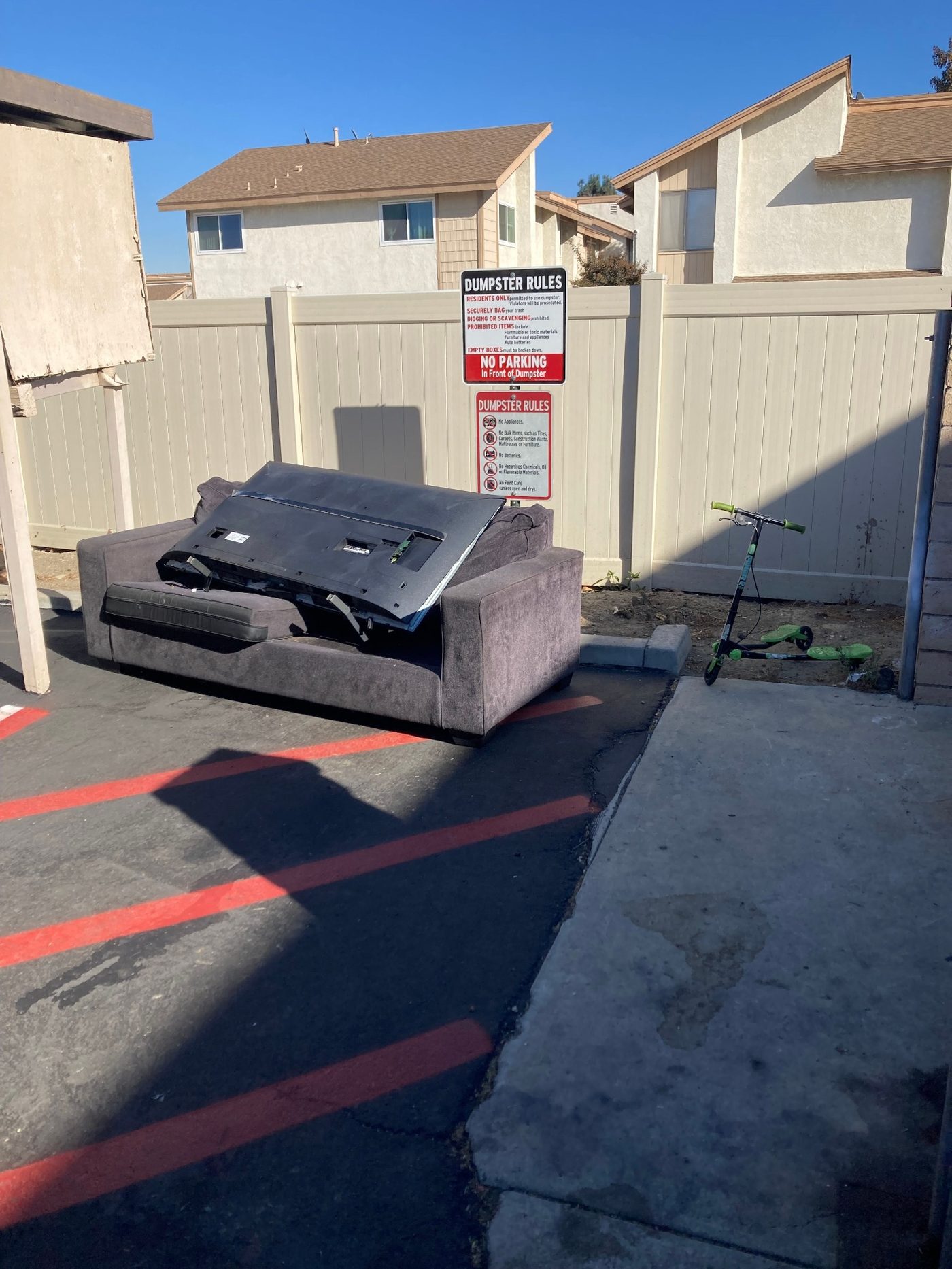 A discarded couch and broken flat-screen television left beside a community dumpster enclosure